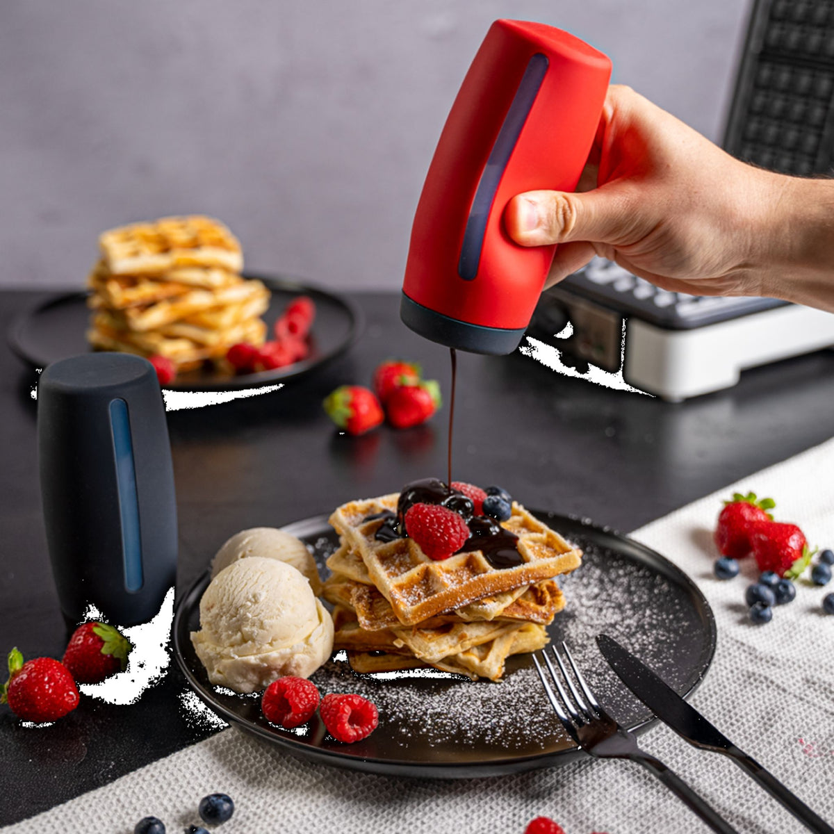 Roter Dosierspender gibt Schokolade über Waffeln mit Beeren und Eis, anthrazitfarbener Spender daneben.