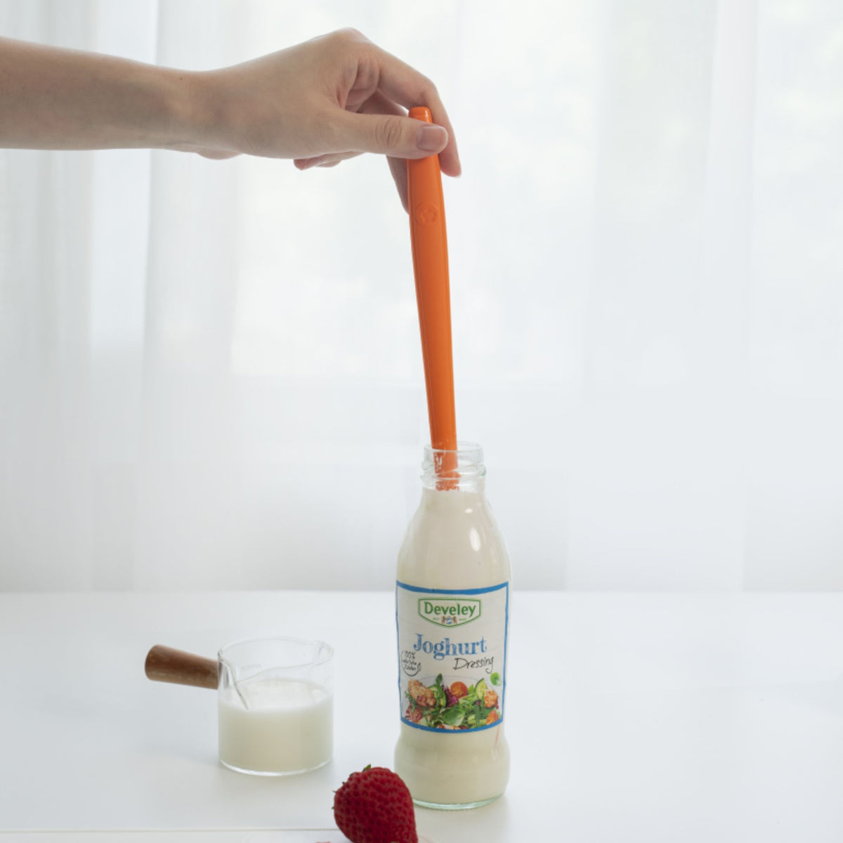 Hand hält orangefarbenen Restelöffel in Glasflasche mit weißer Flüssigkeit.