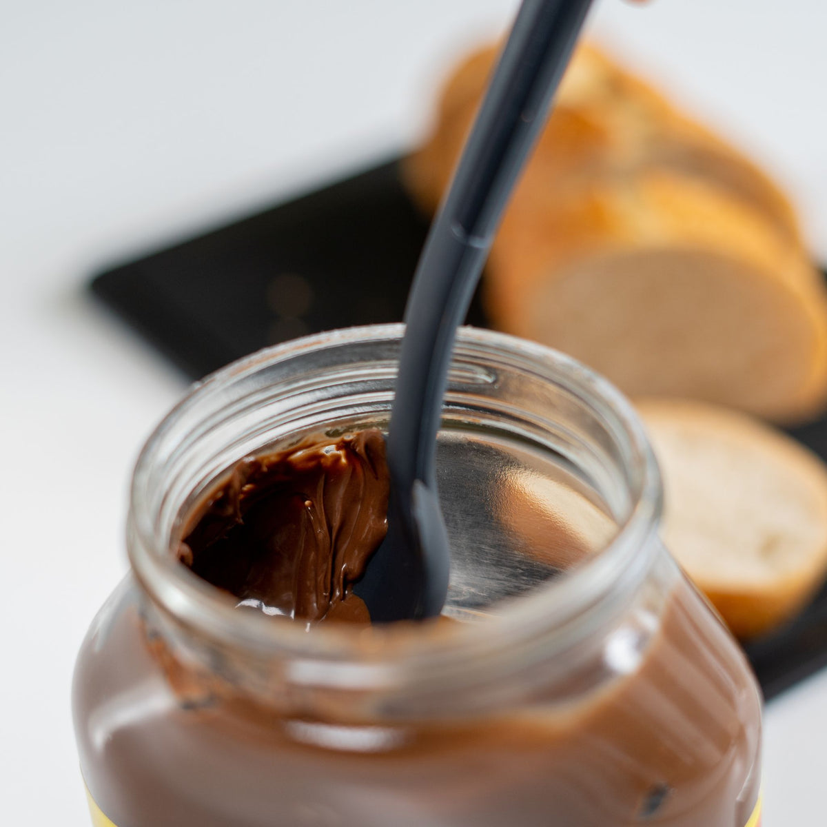 Anthrazitfarbener Restelöffel aus Silikon in Glas mit Schokoaufstrich und Brot im Hintergrund.