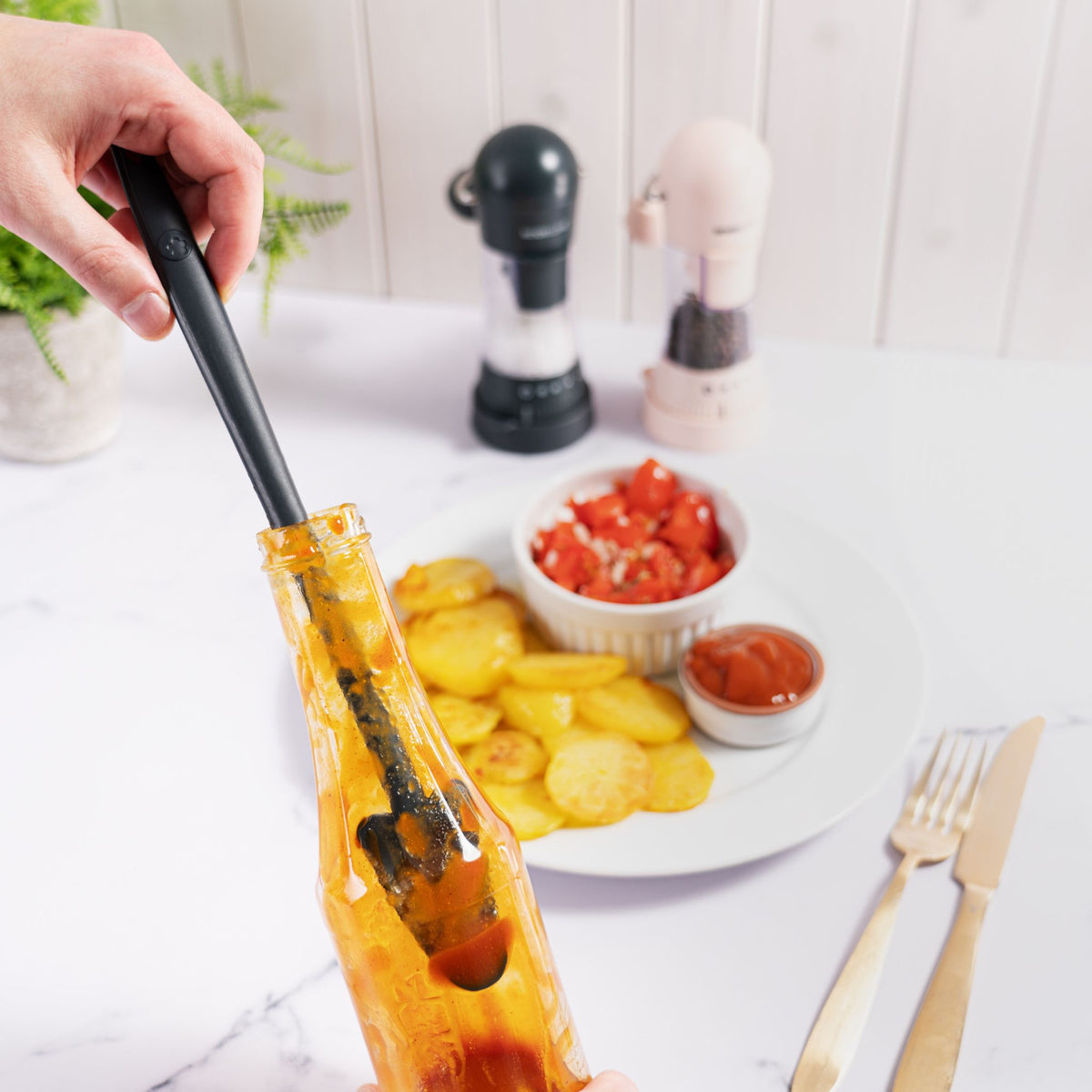 Anthrazitfarbener Restelöffel mit langen Griff, der Sauce von einem Teller mit Kartoffeln aufnimmt.