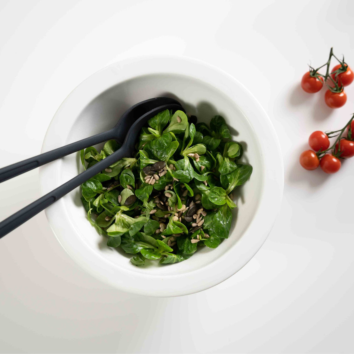 Salatbesteck in Anthrazit liegend in weißer Schüssel mit grünem Salat und Tomatenzweig daneben.