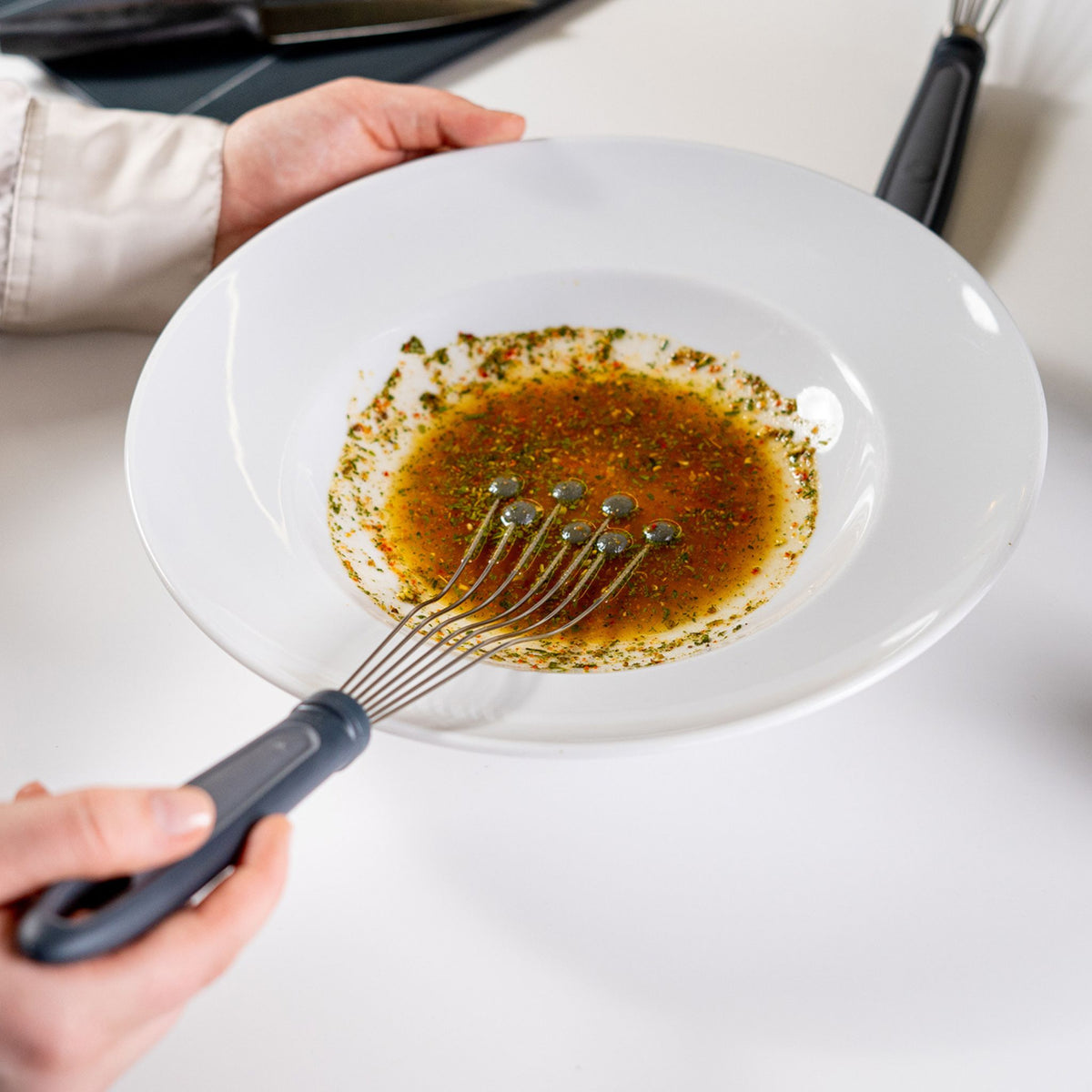 Anthrazitfarbener Tellerbesen mit flexiblem Kopf in weißem Teller mit Dressing.