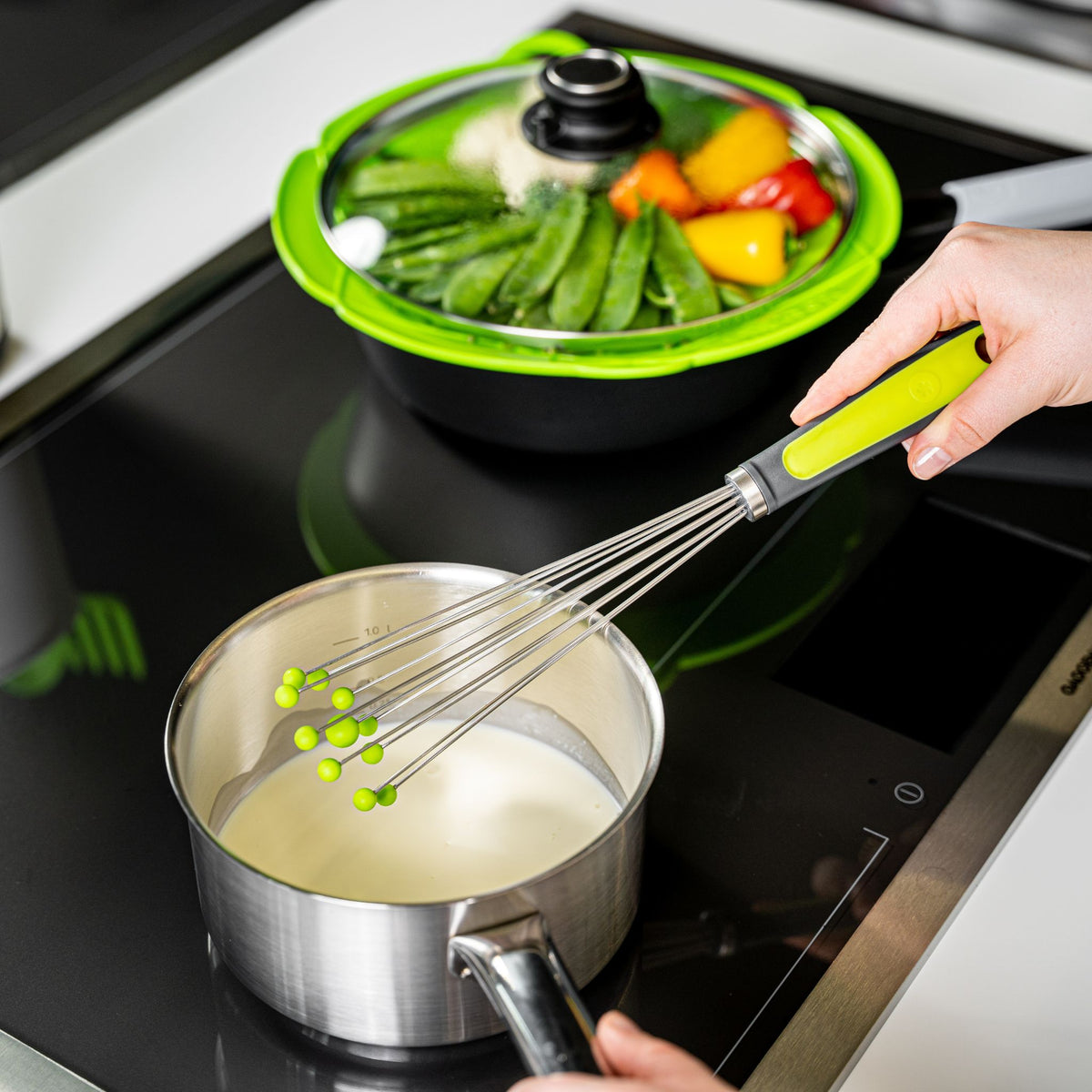 Kugelschneebesen mit limettengrünem Griff, der in einem Topf mit weißer Soße verwendet wird.