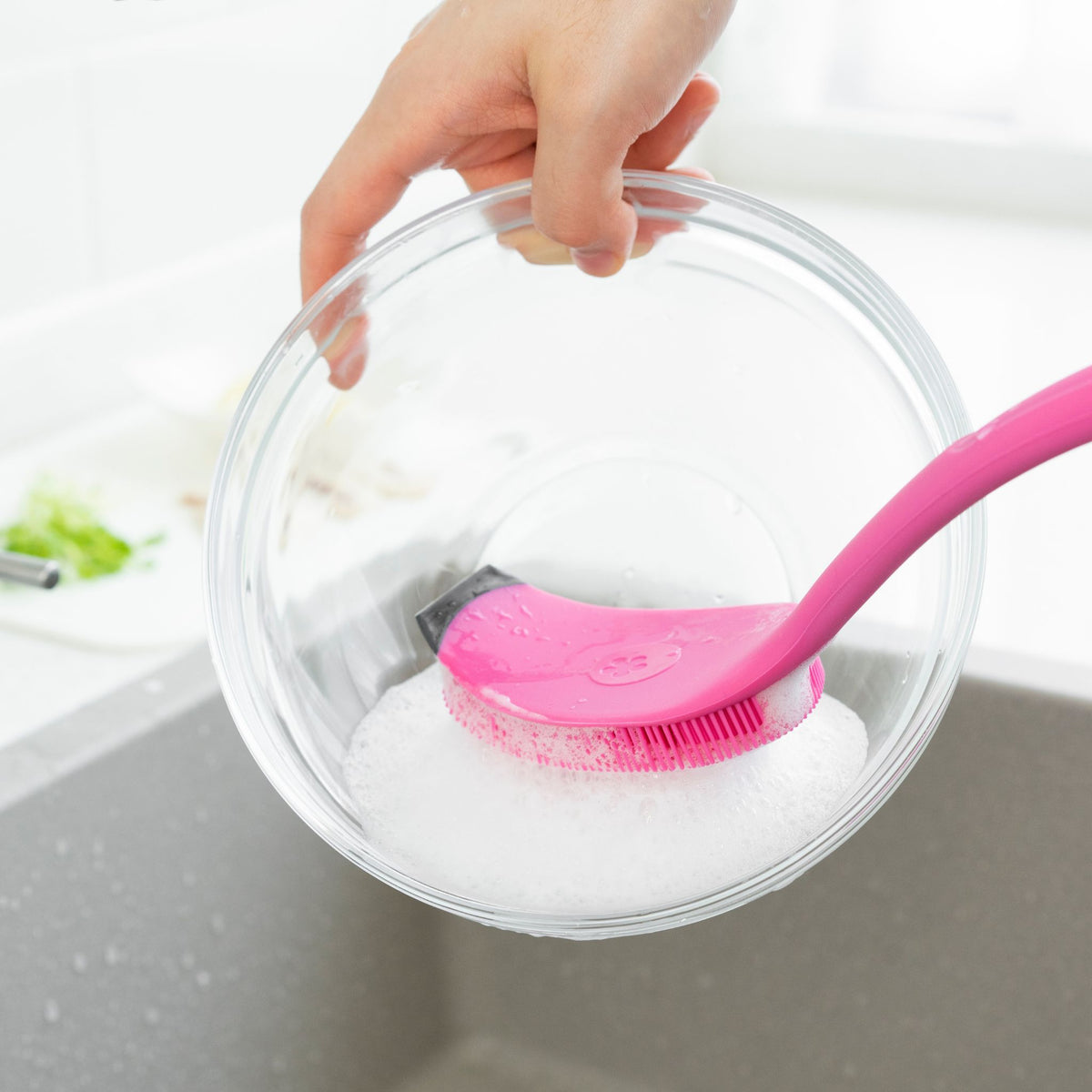 Pinkfarbene Spülbürste mit weichen Silikonborsten reinigt eine Glas-Schüssel über der Spüle.