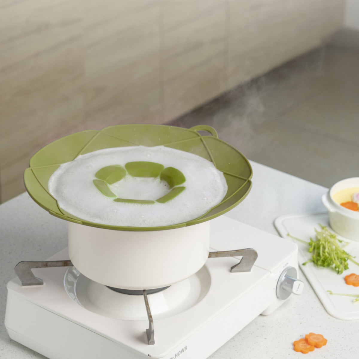 Grüner Silikondeckel mit Blütenform auf weißem Kochtopf vor hellem Hintergrund.