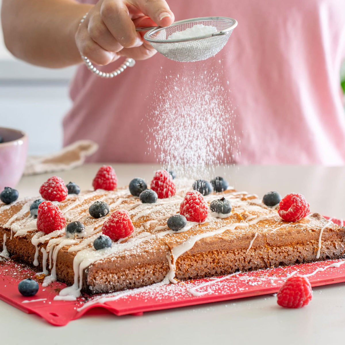 Roter Backrahmen mit Kuchen, mit Beeren und Puderzucker bestreut, Hand siebt Zucker.