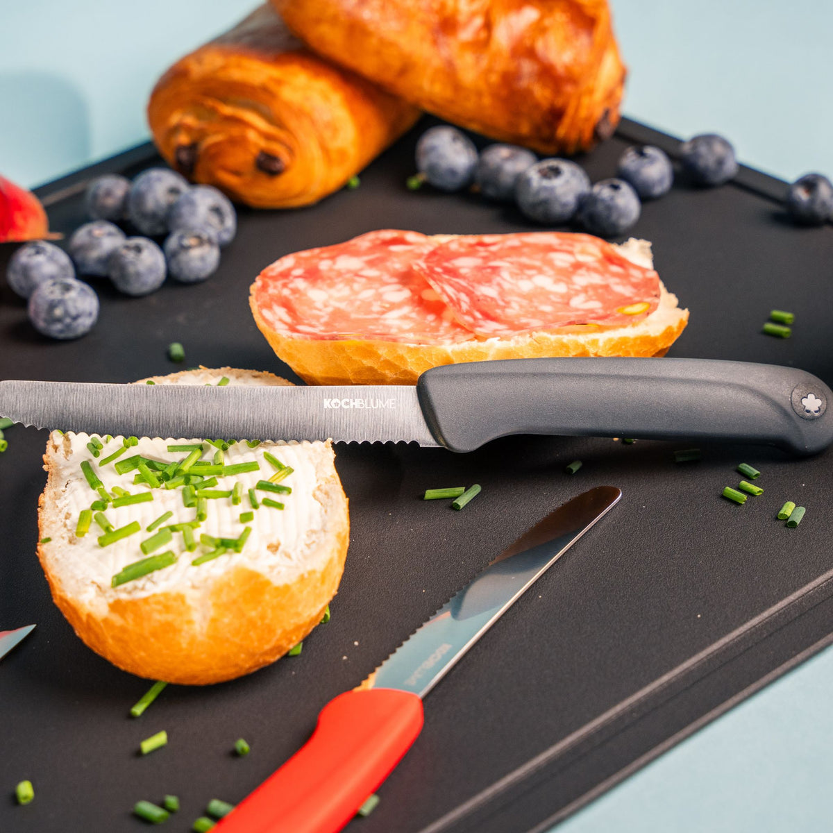 Frühstücksmesser mit schwarzem ergonomischem Griff auf schwarzem Schneidebrett neben Brötchen und Croissants.