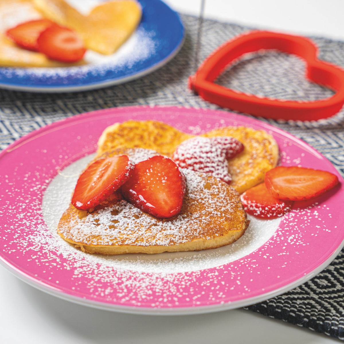 Pfannkuchen in Bärform mit Puderzucker und Erdbeerscheiben auf rosa Teller, roter Herz-Eierformer daneben.