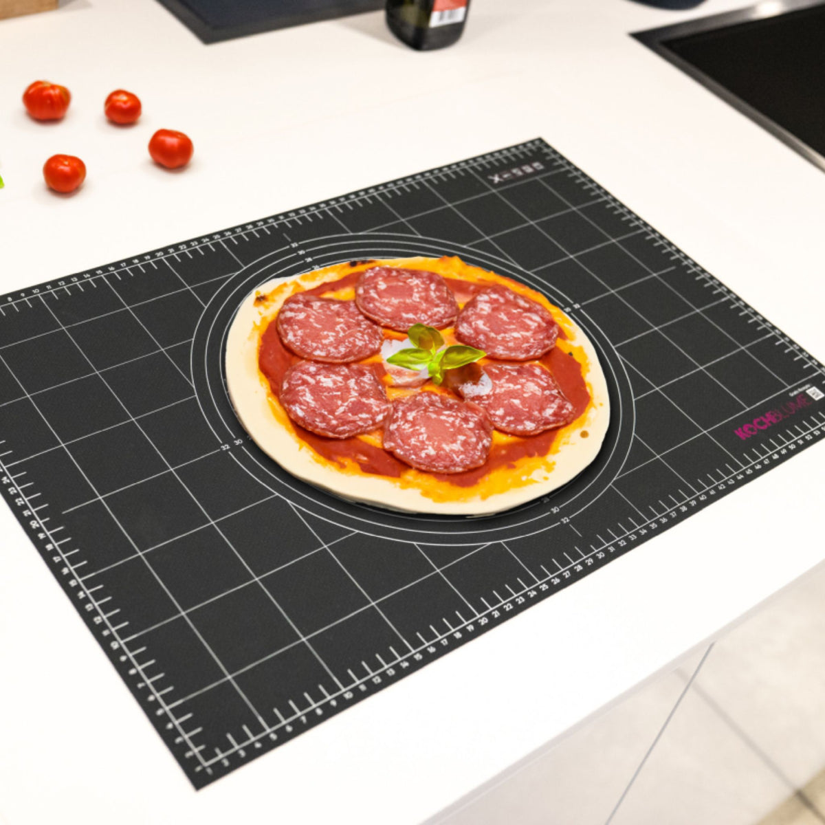Schwarze Gewebematte mit cm-Raster und Pizza mit Salami, auf weißer Küchenarbeitsfläche.